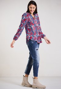 Floral, long-sleeve blouse in blue with red and pink leaves; paired with distressed denim and tan ankle boots. Smooth texture, relaxed fit.