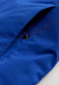 Tessuto blu con superficie texturizzata, dotato di chiusura a patta fissata da un bottone nero. Il materiale appare resistente e liscio.