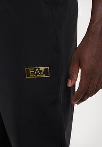 Pantalons de sport noirs avec un logo doré EA7 Emporio Armani sur le côté, texture lisse et design ajusté.