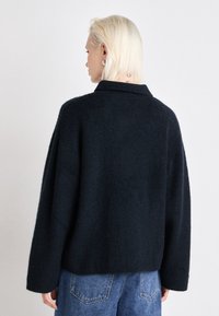 Temno moderna oversize pletena pulover z strukturirano površino, ki ima klasičen ovratnik in spuščena ramenska šiva, v kombinaciji s svetlo modrimi hlačami.