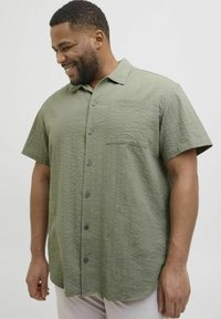 Košulja kratkih rukava s gumbima u prigušenoj maslinastozelenoj teksturiranoj tkanini; sadrži džep na prsima i tamne gumbe.
