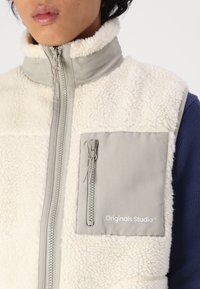 Off-white fleece vest with a high collar, gray zippered pocket, and logo detail. Features a textured surface and contrasting blue material underneath.