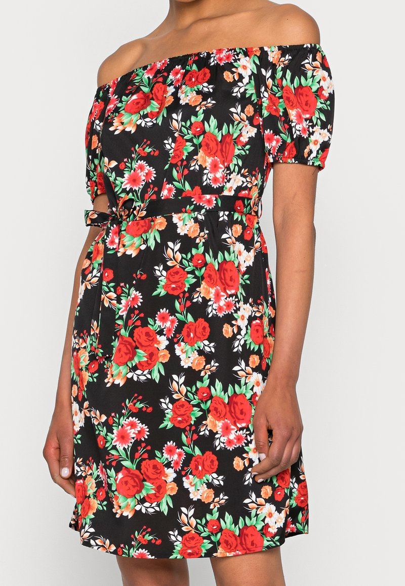 Femme portant une robe noire épaules dénudées avec un motif floral rouge, orange et blanc, des manches courtes bouffantes et une ceinture à nouer à la taille.