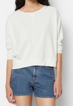 Femme portant un haut blanc à manches longues ample et un short en jean bleu, debout devant un fond blanc uni.