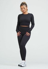 Top corto a coste nero con maniche lunghe abbinato a leggings neri a vita alta. La modella indossa scarpe da ginnastica bianche e posa in modo neutro.