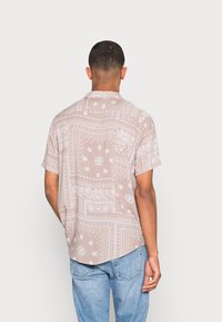Camicia a maniche corte di un beige chiaro con motivi floreali e paisley bianchi, caratterizzata da un orlo dritto e una tasca sul petto.