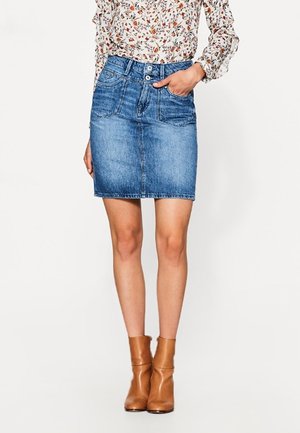 Kobieta ubrana w niebieską mini spódniczkę z denimu, kwiecistą bluzkę z długim rękawem i beżowe botki, stoi z jedną ręką w kieszeni.