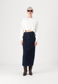 Fehér cropped pulóver, sötétkék farmer maxi szoknya, barna bőrcipők. Egyszerű minták diszkrét varrási részletekkel. Fekete napszemüveg.