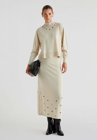 Beige striktrøje med høj krave og dekorerede huller, sat sammen med en matchende beige nederdel. Modellen holder en sort taske og har høje sorte støvler på.