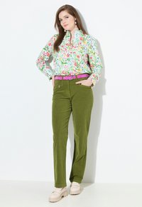 Bluse mit floralem Muster in Weiß mit lebhaften Farben, kombiniert mit olivgrünen Cordhosen und einem rosa Gürtel. Klobige beige Schuhe vervollständigen den Look.
