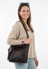 Žena se usmívá, nosí béžový svetr a světlé džíny, má přes rameno černou koženou kabelku a na zápěstí fialkové hodinky, na hlavě sluneční brýle.