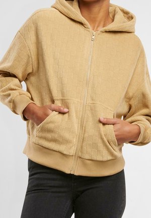 Personne portant un sweat à capuche beige texturé avec fermeture éclair, capuche et poches avant, mains dans les poches, associé à un pantalon foncé.