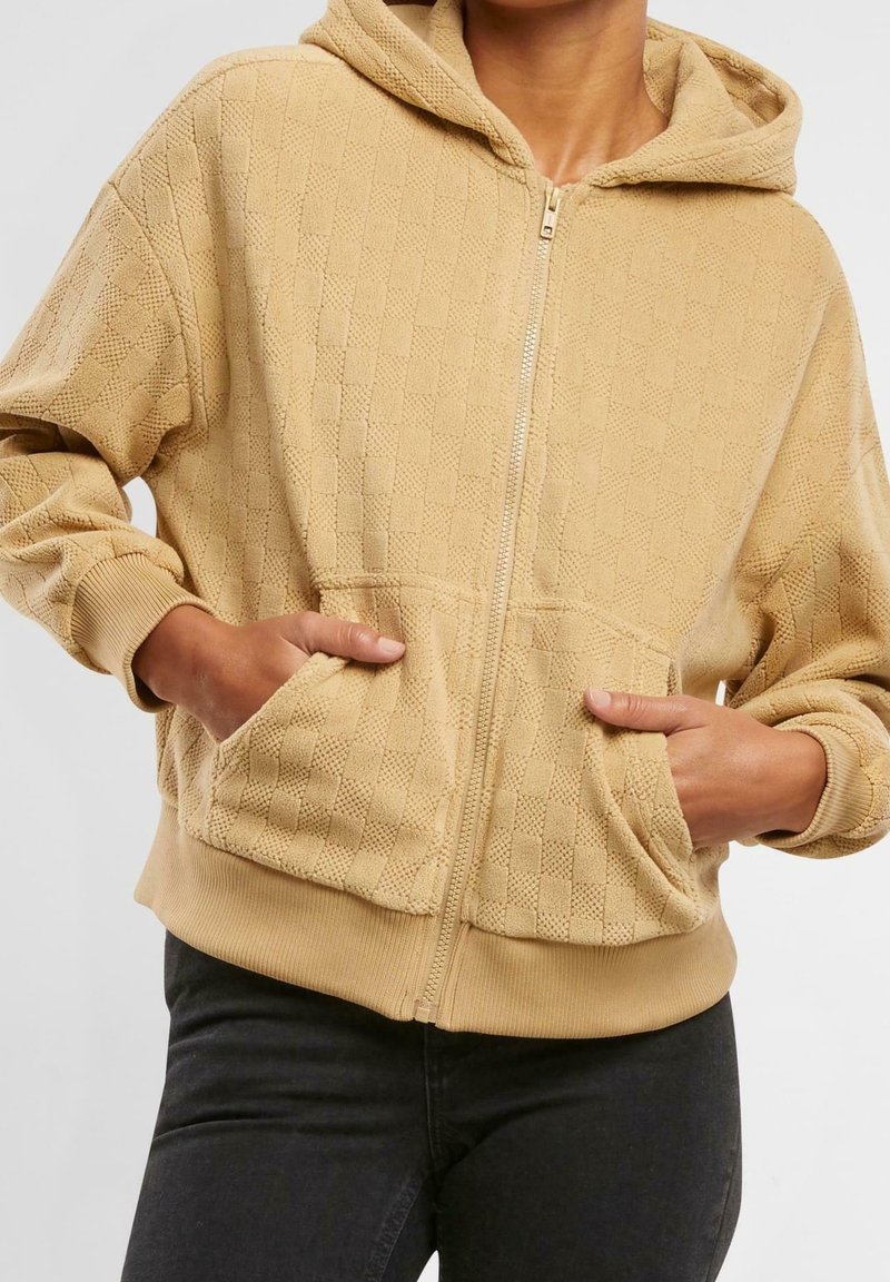 Personne portant un sweat à capuche beige texturé avec fermeture éclair, capuche et poches avant, mains dans les poches, associé à un pantalon foncé.