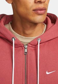 Felpa rossa in cotone con zip, con cordini bianchi e un piccolo logo Nike bianco, dotata di una cerniera argentata e di un rivestimento interno beige.