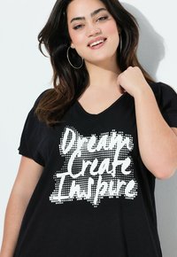 Sort bomulds t-shirt med et hvidt prikket grafisk motiv og teksten "Drøm Skab Inspirer." V-hals design med korte ærmer.