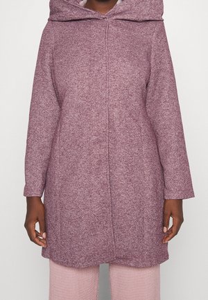 Femme portant un manteau à capuche texturé rose poudré à manches longues, associé à un pantalon rose clair, debout, bras détendus sur les côtés.
