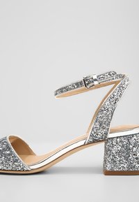 Sandale ouverte à paillettes argentées avec bride à la cheville, talon bloc et semelle intérieure beige sur fond gris clair.