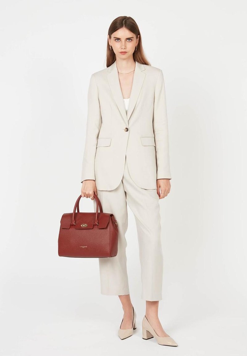 Borsa a mano rossa in pelle con manici arrotolati, finitura testurizzata e hardware dorato. La modella indossa un abito beige chiaro e tacchi alti nudi.