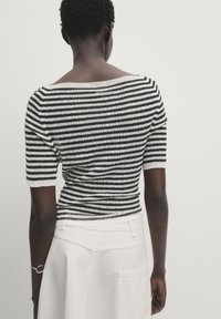 Person med kort, lockigt hår som bär en svartvit randig kortärmad topp och en vit högmidjad kjol, sedd bakifrån.