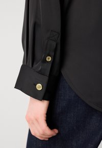 Camisa de cetim preta com botões dourados nos punhos, apresentando um corte ajustado e textura suave. O fundo exibe tecido de ganga escura.