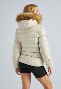 Beige gewatteerde jas met een met bont afgewerkte capuchon, voorzien van horizontale stiknaden en een getailleerd ontwerp. Glad textuur met een glanzende afwerking.