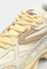 Baskets Lacoste présentant une palette de couleurs crème et beige, en cuir texturé, avec des lacets jaunes et un logo sur la languette et sur le côté.
