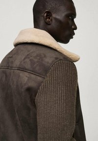 Brun mockaläderjacka med en krämfärgad shearling-krage och texturerade stickade ärmar. Har en figursydd design utan synliga metalldetaljer.