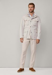 Giacca leggera di un pallido beige con molteplici tasche frontali, colletto alto e bottoni a pressione, abbinata a pantaloni chiari e scarpe marroni.