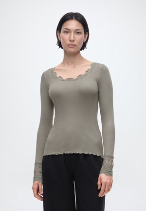 Frau mit kurzen dunklen Haaren, die ein eng anliegendes, geripptes, olivgrünes Langarmshirt mit Spitzenverzierung am Ausschnitt und an den Bündchen sowie schwarze Hosen trägt.