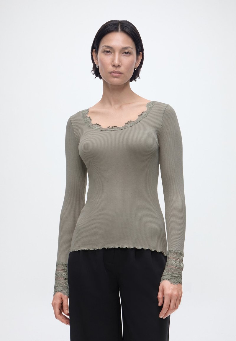 Γυναίκα με κοντά σκούρα μαλλιά που φοράει εφαρμοστή ριμπ πλεκτή μπλούζα μακρυμάνικη σε ελιά πράσινο χρώμα με δαντέλα στο ντεκολτέ και τα μανίκια, και μαύρο παντελόνι.