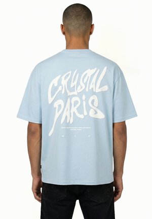 Uomo che indossa una maglietta azzurro chiaro con grande scritta bianca "CRYSTAL PARIS" sulla schiena, abbinata a pantaloni neri, con la schiena rivolta verso la fotocamera.