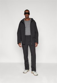 Veste imperméable noire avec capuche, pull gris, pantalon noir et baskets claires. Modèle portant de grandes lunettes de soleil sombres.