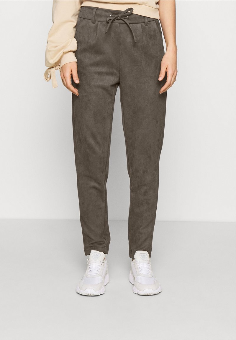 Personne portant un pantalon droit gris foncé avec un cordon à l'avant et des baskets blanches, debout devant un fond clair uni.