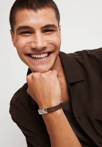 Pulsera de cuero marrón con detalle de herrajes plateados, llevada en una mano apoyada en la barbilla. Modelo con camisa oscura abotonada.