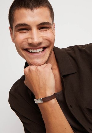 Pulsera de cuero marrón con detalle de herrajes plateados, llevada en una mano apoyada en la barbilla. Modelo con camisa oscura abotonada.