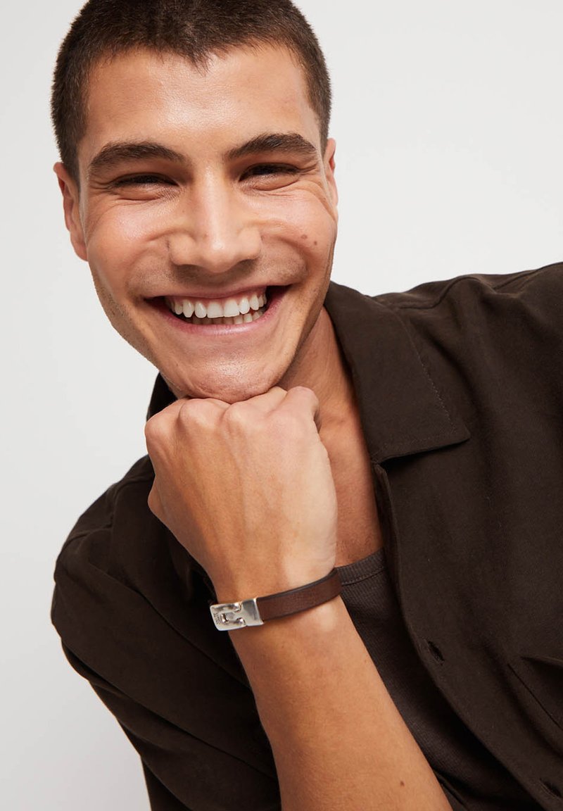 Pulsera de cuero marrón con detalle de herrajes plateados, llevada en una mano apoyada en la barbilla. Modelo con camisa oscura abotonada.