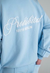 Helles blaues Sweatshirt mit weißem besticktem Text "Prohibited 10119 Berlin." Verfügt über gerippte Bündchen und einen lockeren Schnitt. Weiche Stoffbeschaffenheit.