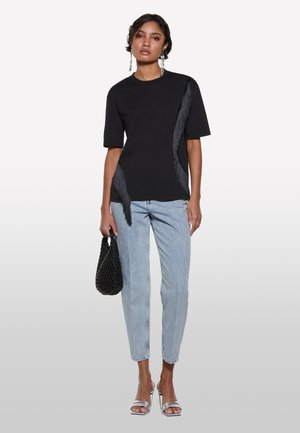 T-shirt nero a maniche corte con dettagli di frange testurizzati su un lato, abbinato a jeans blu chiaro e una borsa a tracolla nera intrecciata. Sandali argento.