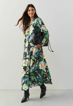 Langes Blumenkleid mit schwarzem Hintergrund, bunten Blüten und Punkten, kombiniert mit einer schwarzen Beuteltasche und spitzen schwarzen Stiefeln.