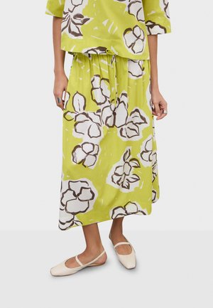 Woman wearing green floral midi skirt and matching top with cream-colored flat Mary Jane shoes, standing against plain background.