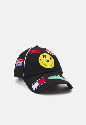 Casquette de baseball noire avec des patches brodés colorés comprenant un smiley jaune avec "NY", "1985", logos Tommy Jeans et des accents rouges, blancs et verts.