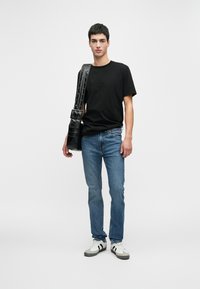 Jovem em pé, vestindo uma t-shirt preta, jeans azuis, ténis brancos e a carregar uma mala de ombro de couro preta com alça larga.