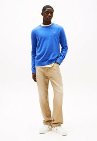 Maglione blu a maglia con polsini e orlo a costine, indossato sopra una camicia gialla, abbinato a pantaloni beige a gamba dritta e sneaker bianche.