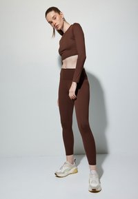 Brun langærmet crop top parret med matchende højtaljede leggings, begge lavet af glat stof. Model iført beige træningssko.