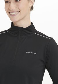 Sort quarter-zip atletisk top lavet af strækstof. Har hvide detaljer på skuldrene og "ENDURANCE" logoet på brystet. Glat tekstur.