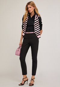 Pantalones ajustados negros combinados con una chaqueta sin mangas a rayas en rosa y negro sobre una camisa negra. Accesorios incluyen un bolso rosa y tacones negros.