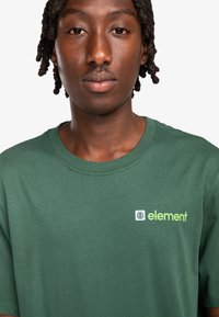 Camiseta de algodón verde con cuello redondo, que presenta un pequeño logo en blanco y verde claro en la zona del pecho izquierdo. Textura suave.