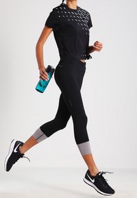 Mustat urheilulliset crop-topit, joissa on heijastava kuvio, yhdistettyinä mustiin legginsseihin, joissa on harmaita yksityiskohtia. Mustat juoksukengät, pitäen kädessä tealli vedenpullon.