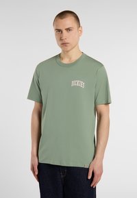 Zelené krátké bavlněné tri čko s kulatým výstřihem a vyšitým logem "Dickies" v růžové barvě na levém prsu. Model má na sobě tmavé džíny.