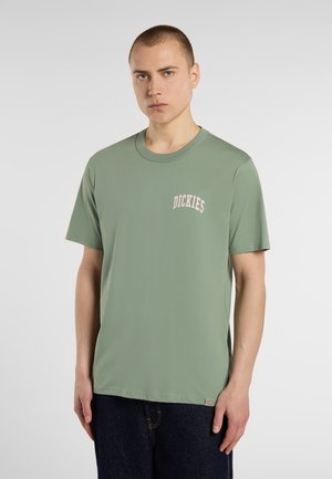 Green short-sleeve cotton T-shirt with a crew neck and embroidered "Dickies" logo in pink on the left chest. Model wears dark denim jeans.